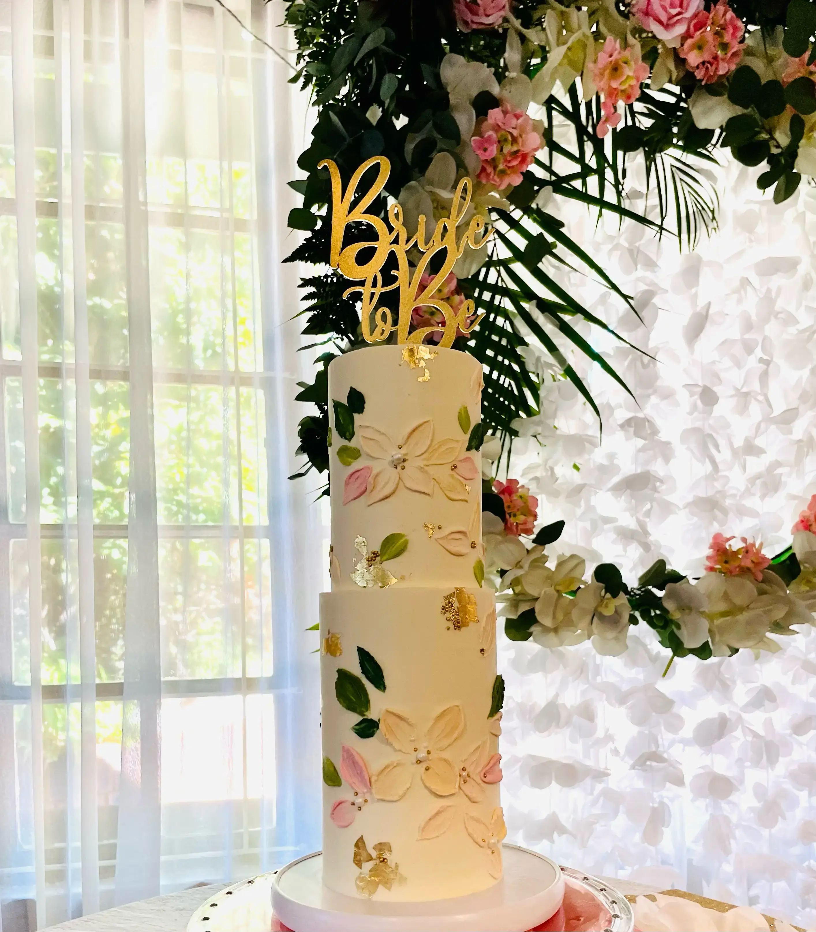 Two-tier cream cake with hand-painted floral detail and gold leaf accents, “Bride to Be” topper