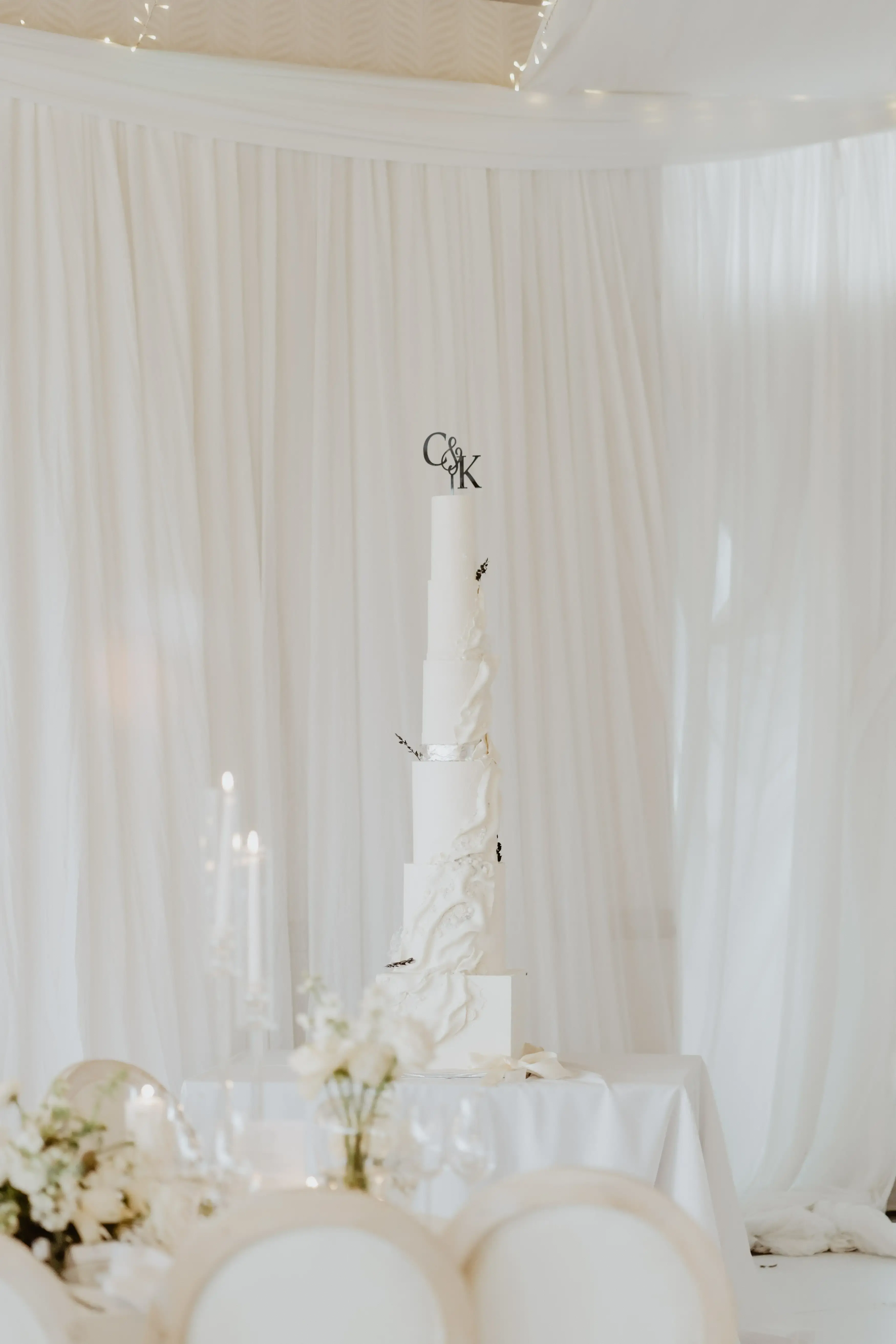 Tall white wedding cake with a sculpted torn-paper texture on the lower tiers and a monogram topper, photographed with candles against a sheer drapery backdrop