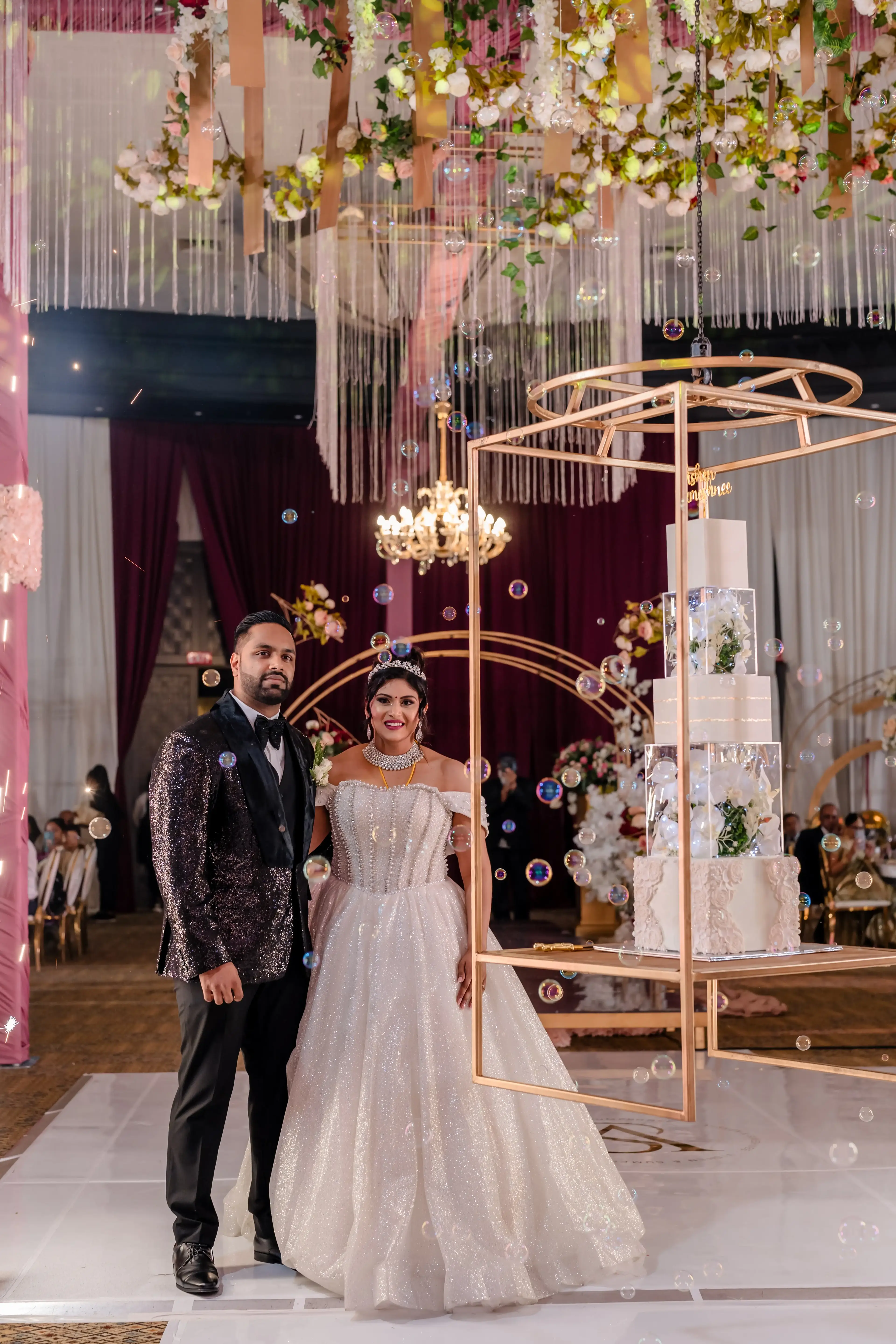 Bride and groom beside a four-tier wedding cake displayed in a hanging gold frame with floating bubbles