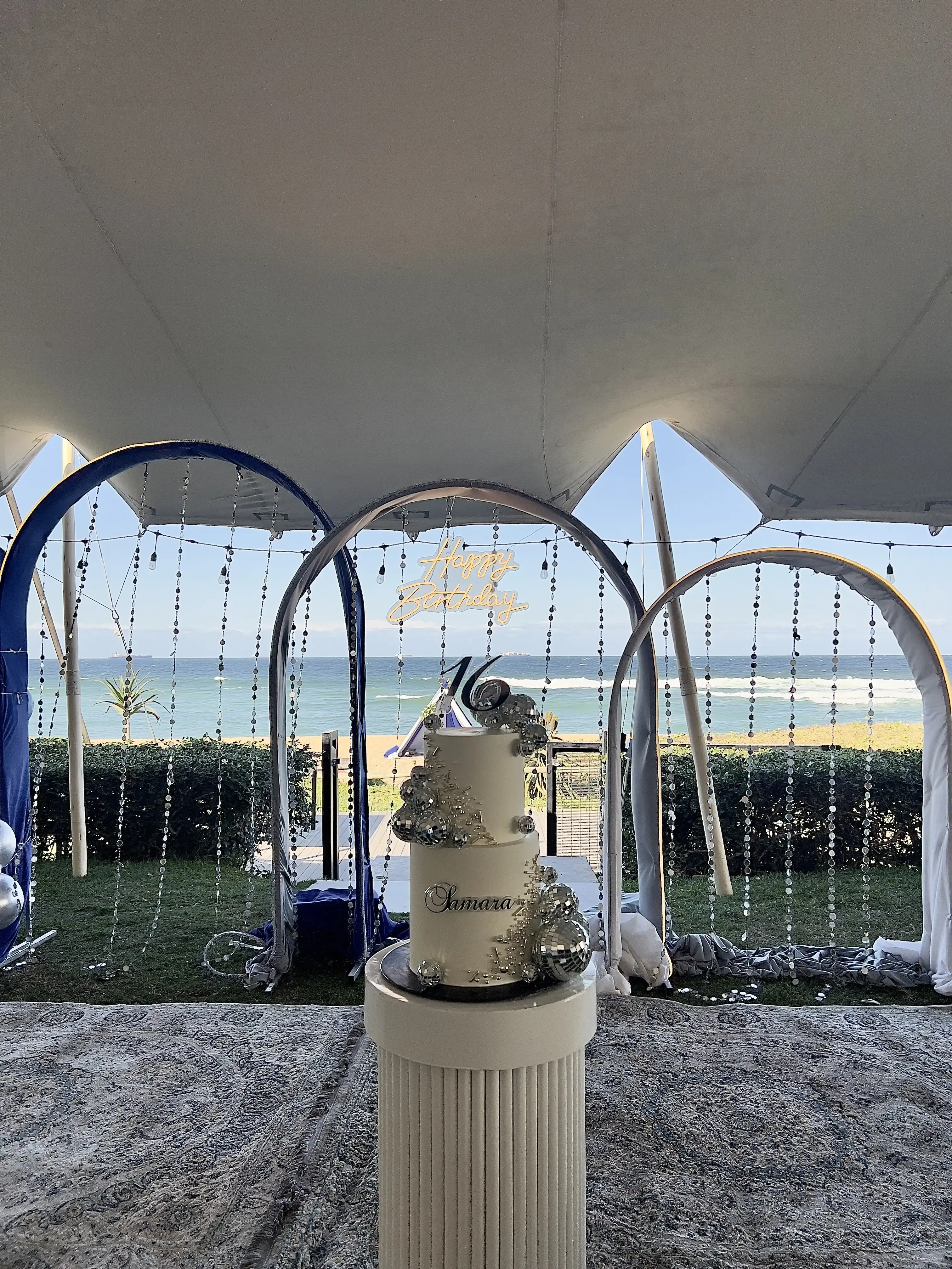 White two-tier cake with silver disco-ball clusters and a “16” topper, set under a beachside marquee