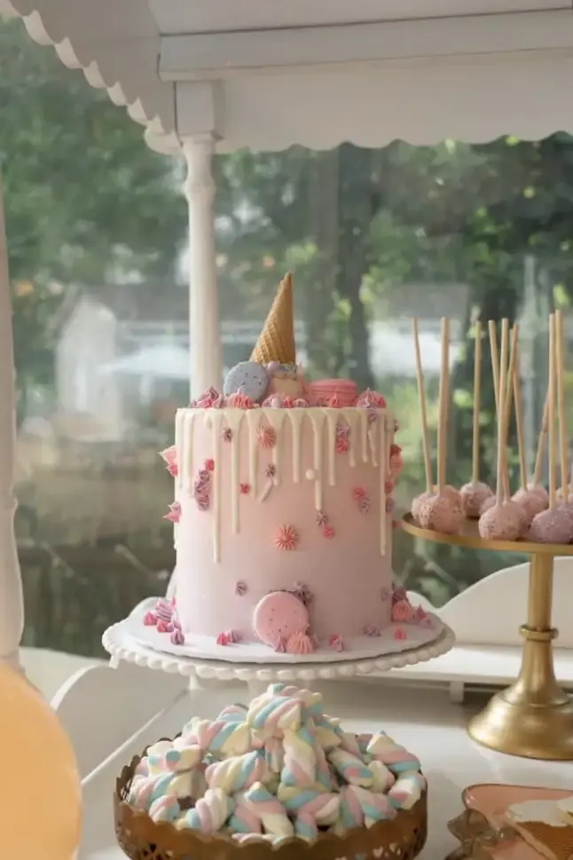Pink drip cake with an ice cream cone topper and macarons, next to a stand of sprinkle cake pops