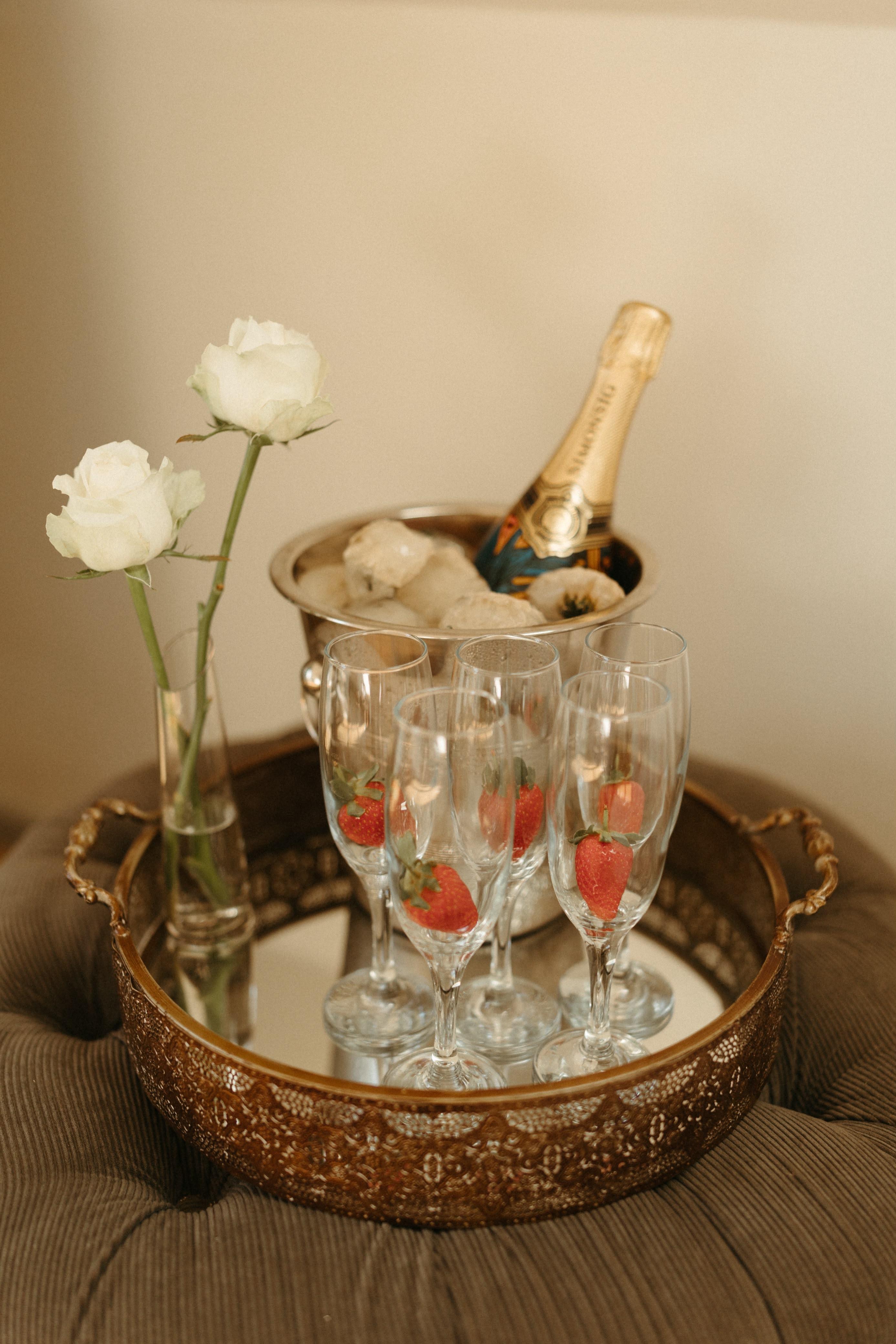 A small styled tray with white roses, a bottle of sparkling wine on ice and strawberry-decorated champagne flutes