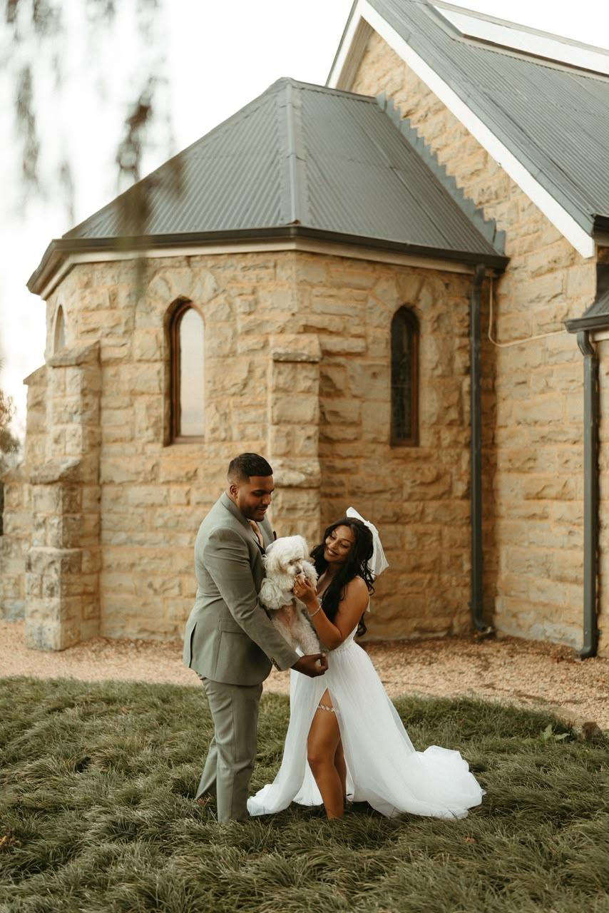 A couple outside a stone chapel with a small dog, one of them kneeling in a full tulle skirt
